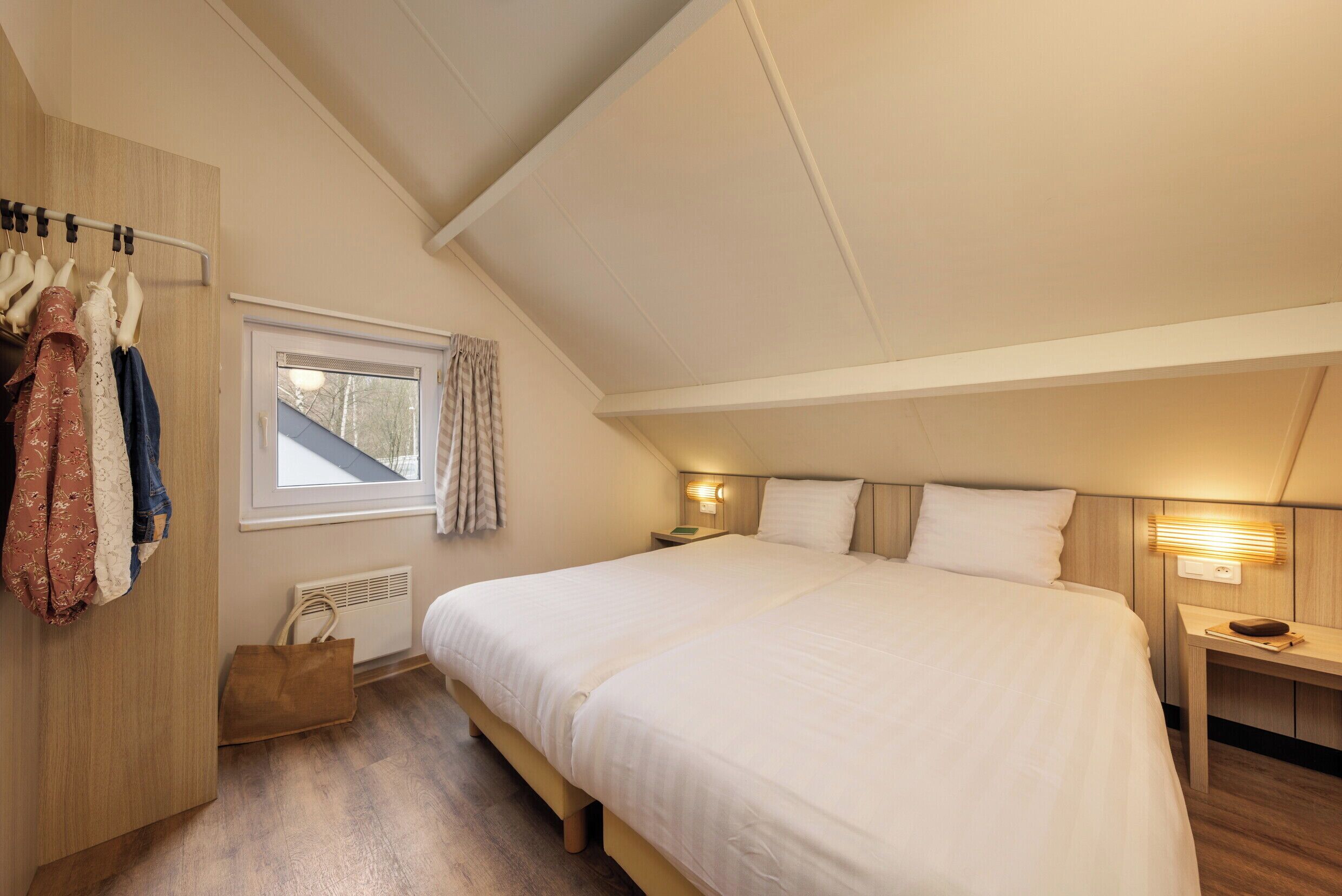 Een slaapkamer met een groot bed, een garderobe, een raam met gordijnen, een nachtkastje met een lamp en een houten vloer.