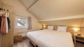 Een slaapkamer met een groot bed, een garderobe, een raam met gordijnen, een nachtkastje met een lamp en een houten vloer.
