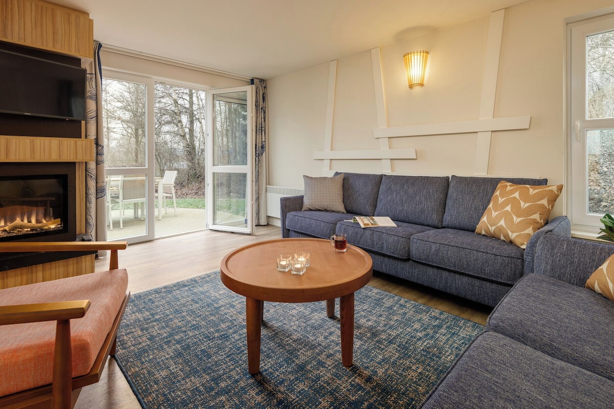 Un salon moderne avec une cheminée, un canapé, une table basse et une grande fenêtre donnant sur la forêt.