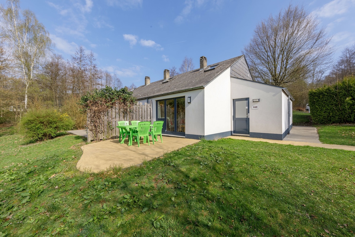 A small, single-story house with a number 140 on the door, a patio with green chairs, and a garden with trees and shrubs.