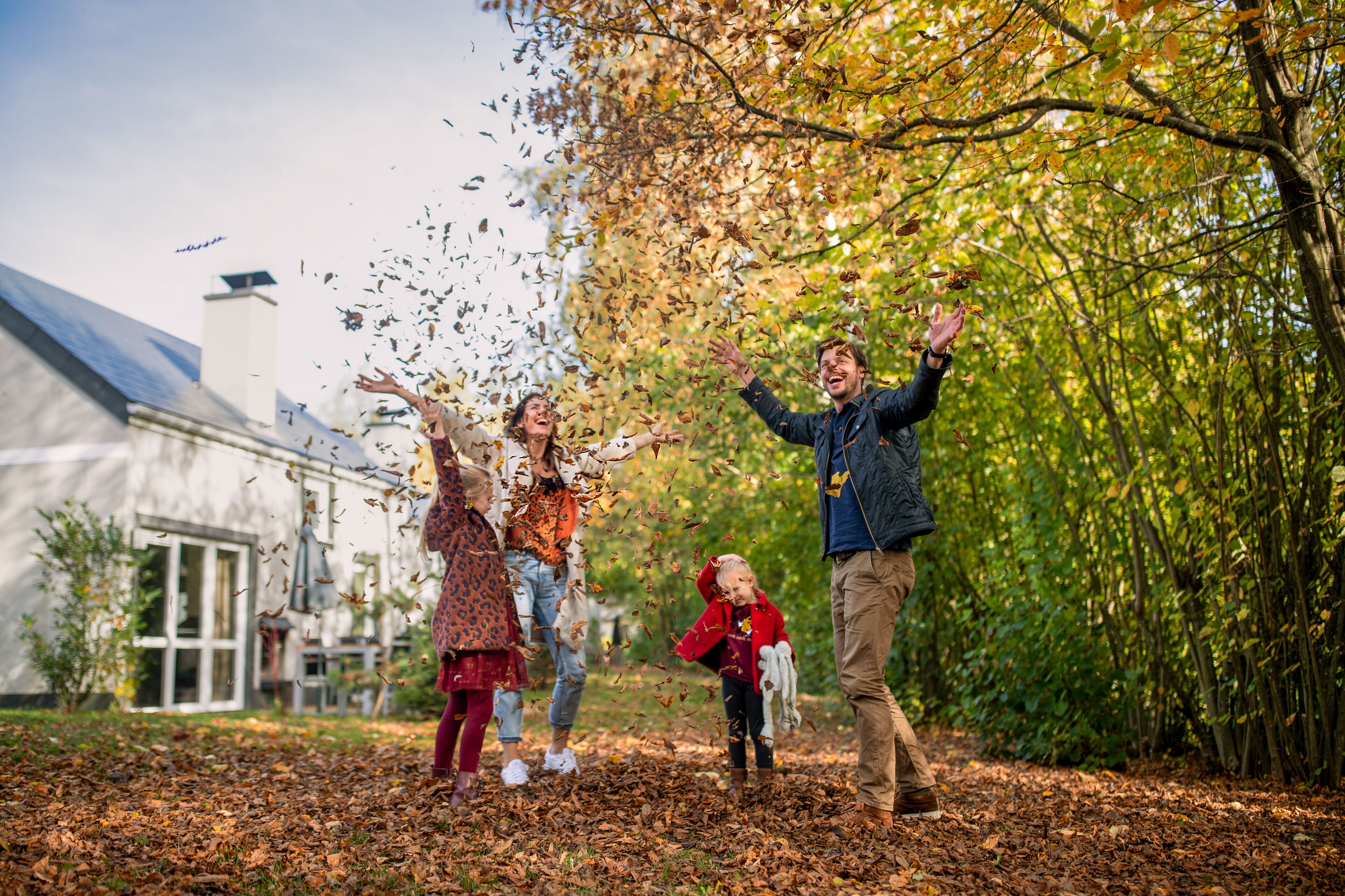 Une famille joue avec des feuilles dans un parc, avec une maison en arrière-plan.
