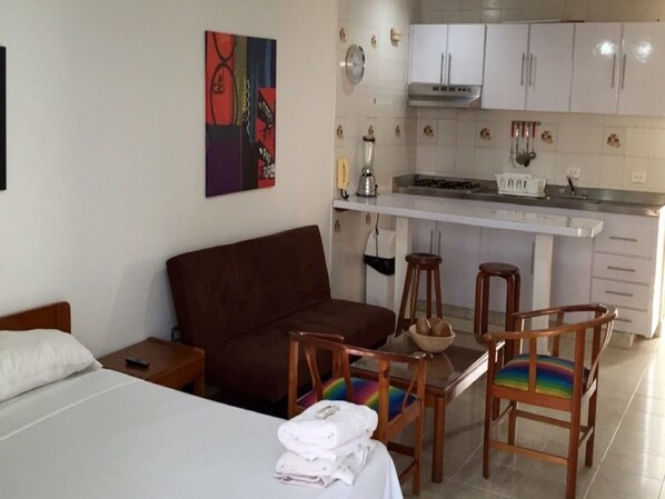 Una habitación de hotel con una cama, un sofá marrón, una pequeña mesa con sillas, un área de cocina con armarios blancos y una estufa, y un cuadro abstracto colorido en la pared.