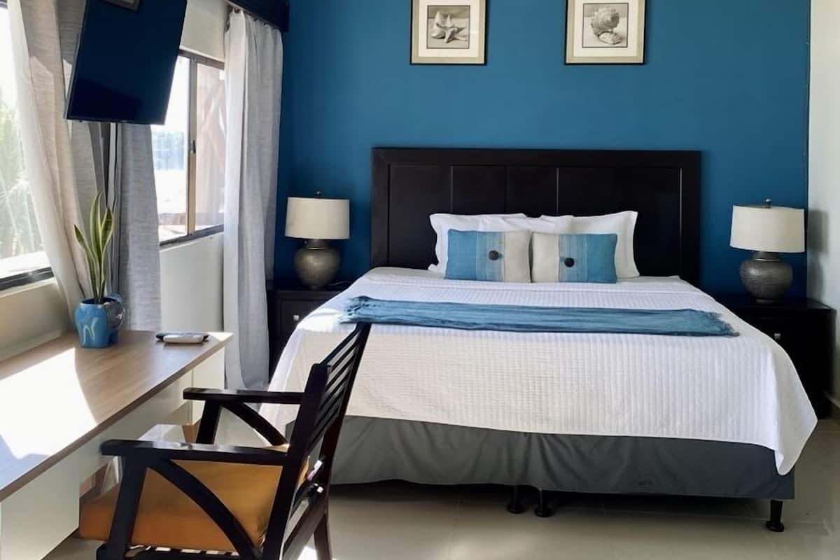 A bedroom with a bed, two bedside tables with lamps, a desk with a chair, a potted plant, and two framed pictures on the wall.