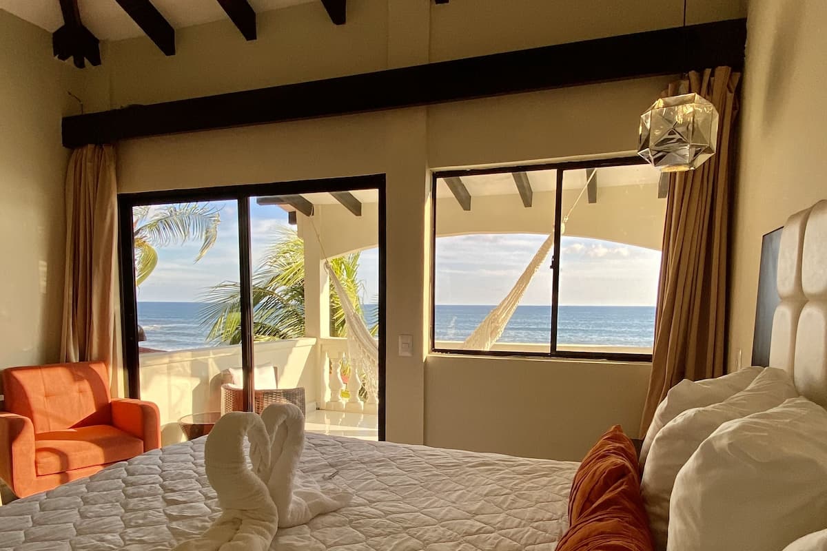 A bedroom with a large bed, a view of the ocean, and a balcony with a chair.