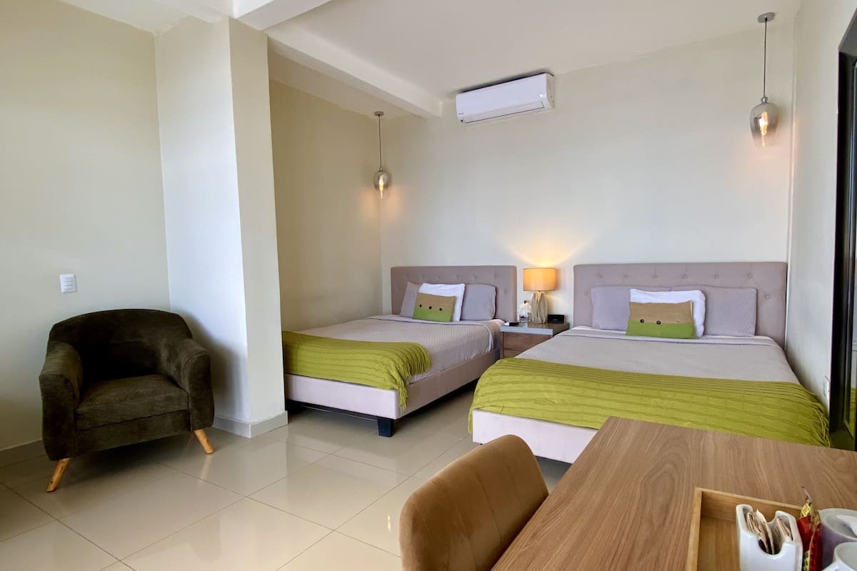 A hotel room with two beds, a wooden table, a chair, and a small armchair.
