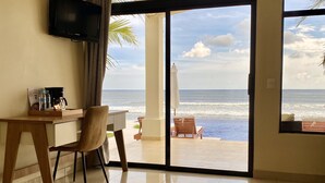 A hotel room with a desk, chair, and two beds. There is a TV on the wall and a view of the ocean through the large sliding glass door.