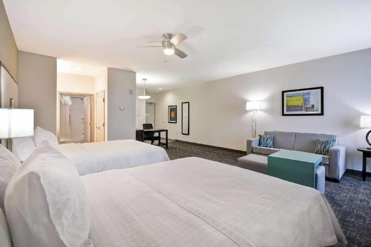 Une chambre d’hôtel avec deux lits, un bureau, un canapé, un ventilateur de plafond et un tableau encadré au mur.