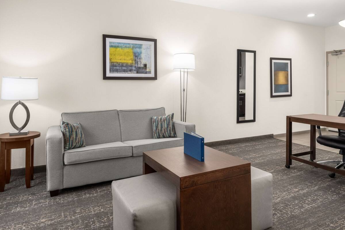 Une chambre d’hôtel moderne avec un canapé gris, une table basse en bois, une petite table d’appoint et un bureau avec une chaise. Deux œuvres d’art encadrées ornent le mur, et une lampe sur pied est placée dans un coin de la pièce.