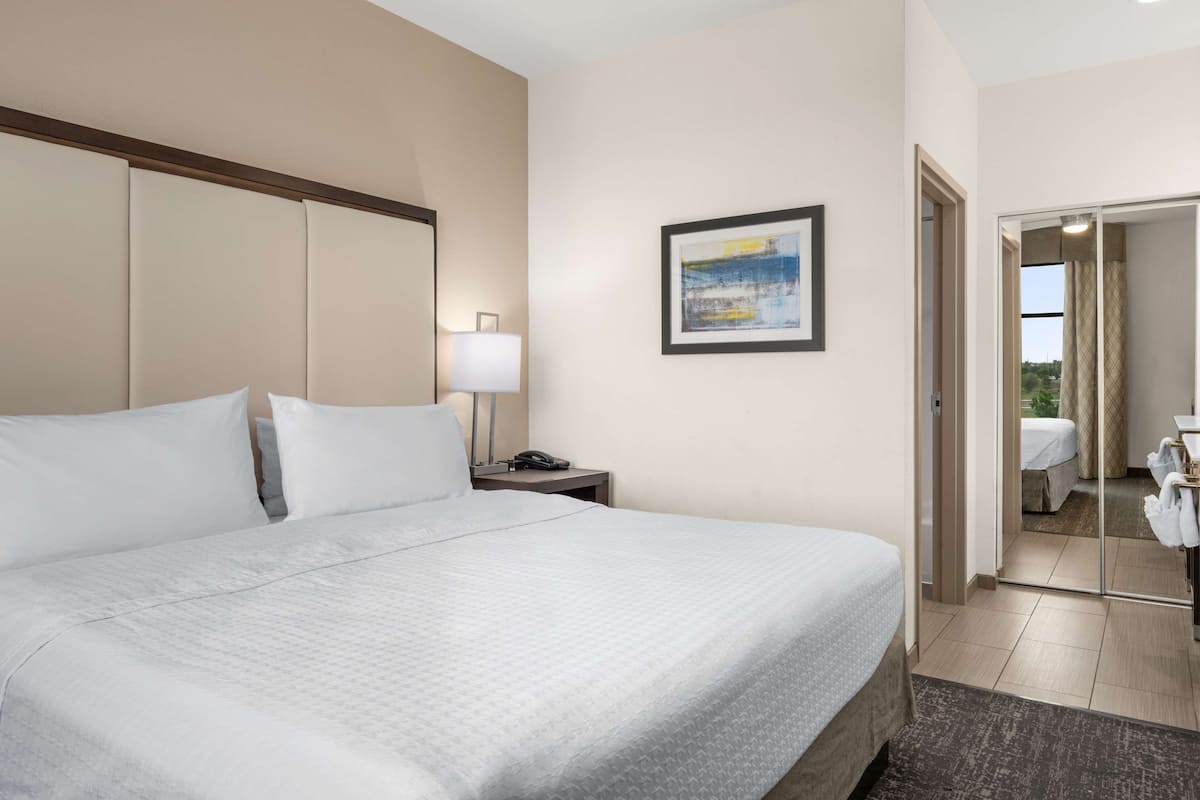 Une chambre d’hôtel avec un grand lit, une table de chevet avec une lampe, un tableau encadré au mur et une salle de bain visible à travers une porte ouverte.