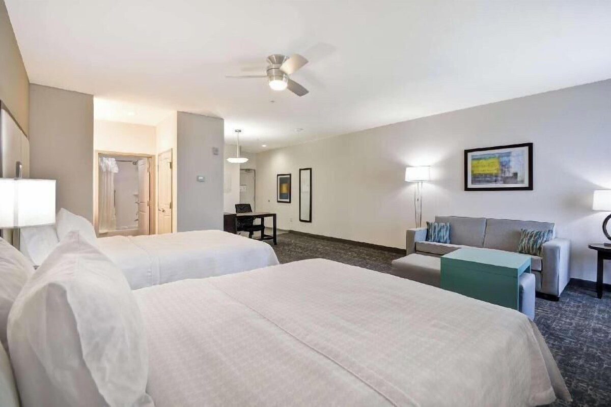 Habitación de hotel con dos camas, un escritorio, un sofá, un ventilador de techo y un cuadro en la pared.