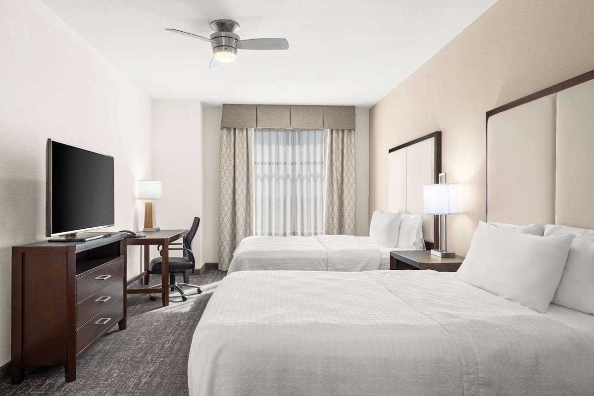 Une chambre d’hôtel avec deux lits, un bureau avec une télévision, une chaise, une lampe de bureau et un ventilateur de plafond.