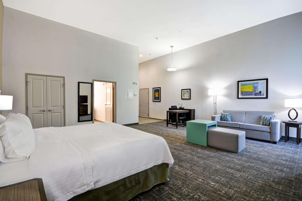 Habitación de hotel con una cama grande, un sofá, un escritorio y una silla. Se ven puertas que conducen a otras habitaciones y a un pasillo.