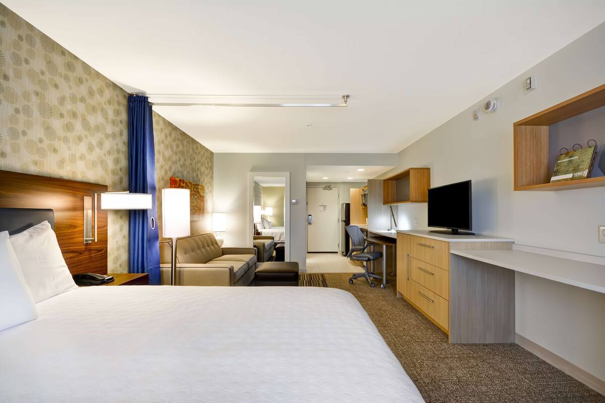 A hotel room with a large bed, a desk with a TV, a chair, and a small kitchenette.