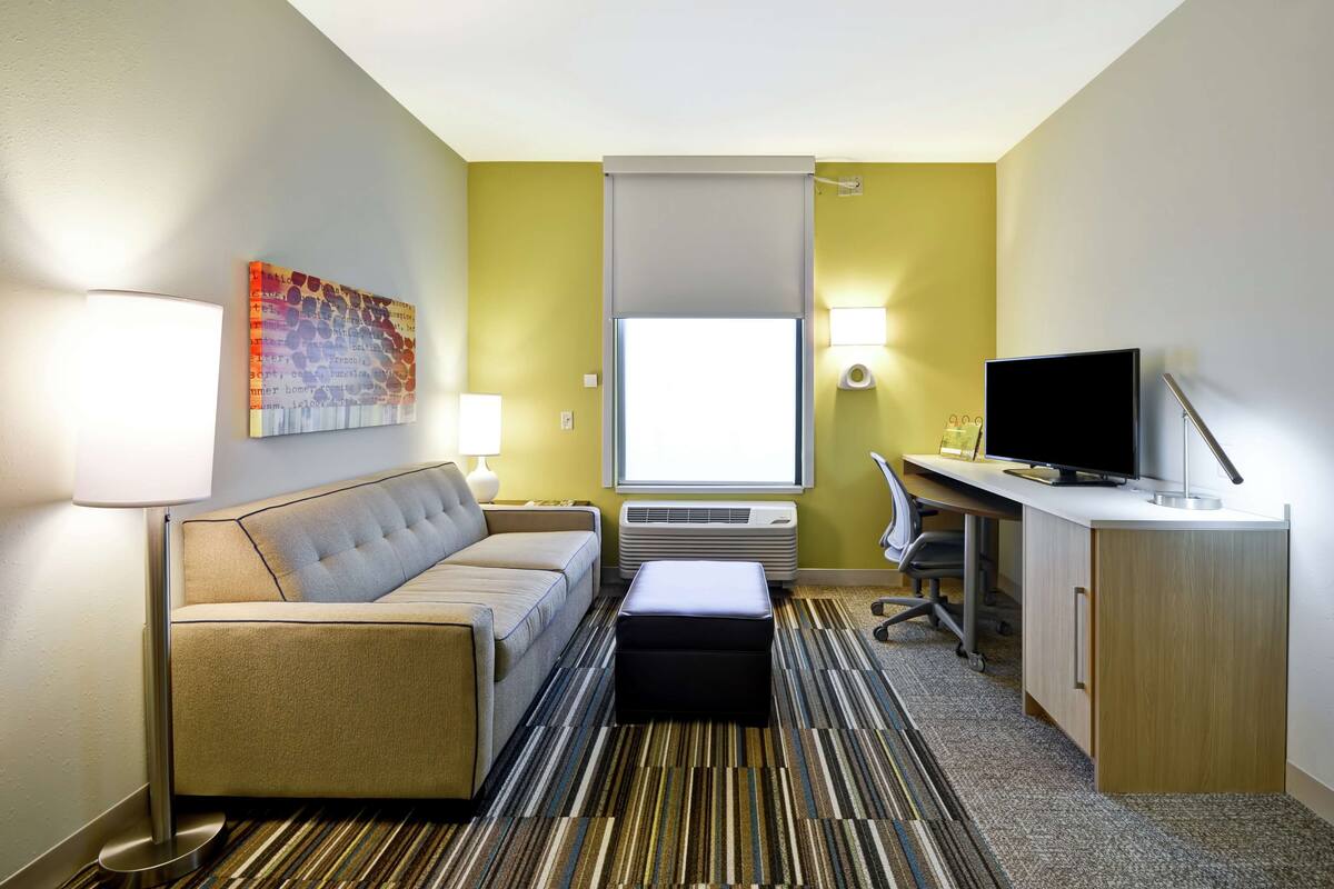 A hotel room with a sofa, a desk, a television, a lamp, an air conditioner, and a window with a view.
