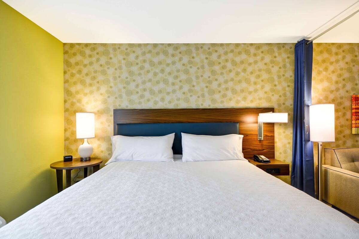 A hotel room with a large bed, two bedside tables, a nightstand, a lamp, and a curtain.