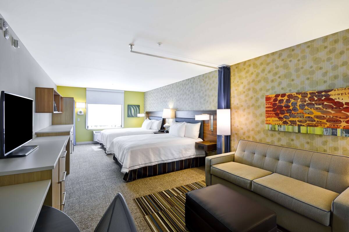A hotel room with a bed, two bedside tables, a sofa, a desk with a flat-screen TV, and a large artwork on the wall.