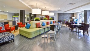 A modern hotel lobby with a green sofa, red and blue pillows, a coffee bar, and a dining area with white chairs and a table. - Home2 Suites by Hilton Decatur Ingalls Harbor (Decatur)