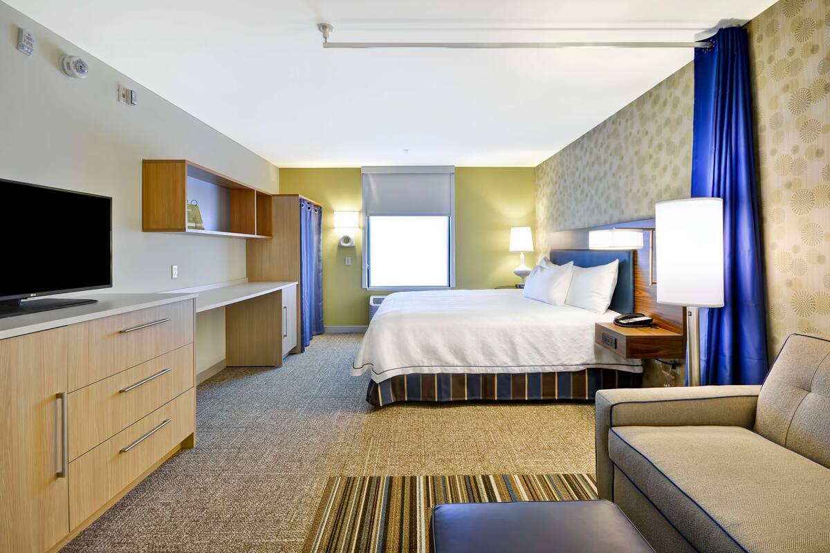 A hotel room with a bed, a TV, a desk, a sofa, and a window with curtains.