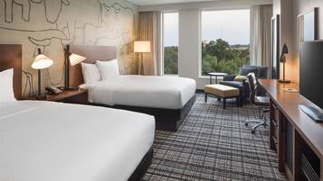 A hotel room with two beds, a desk, a chair, a TV, and a view of the outdoors.
