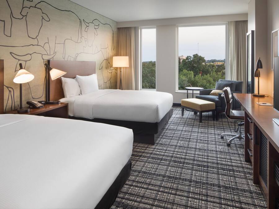 A hotel room with two beds, a desk, a chair, a TV, and a view of the outdoors.