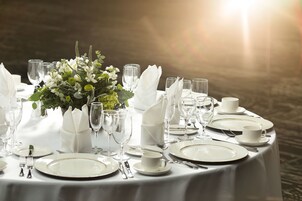 Mesa redonda dispuesta para una comida formal, con mantel blanco, platos, cubiertos, copas y un centro de mesa con flores.