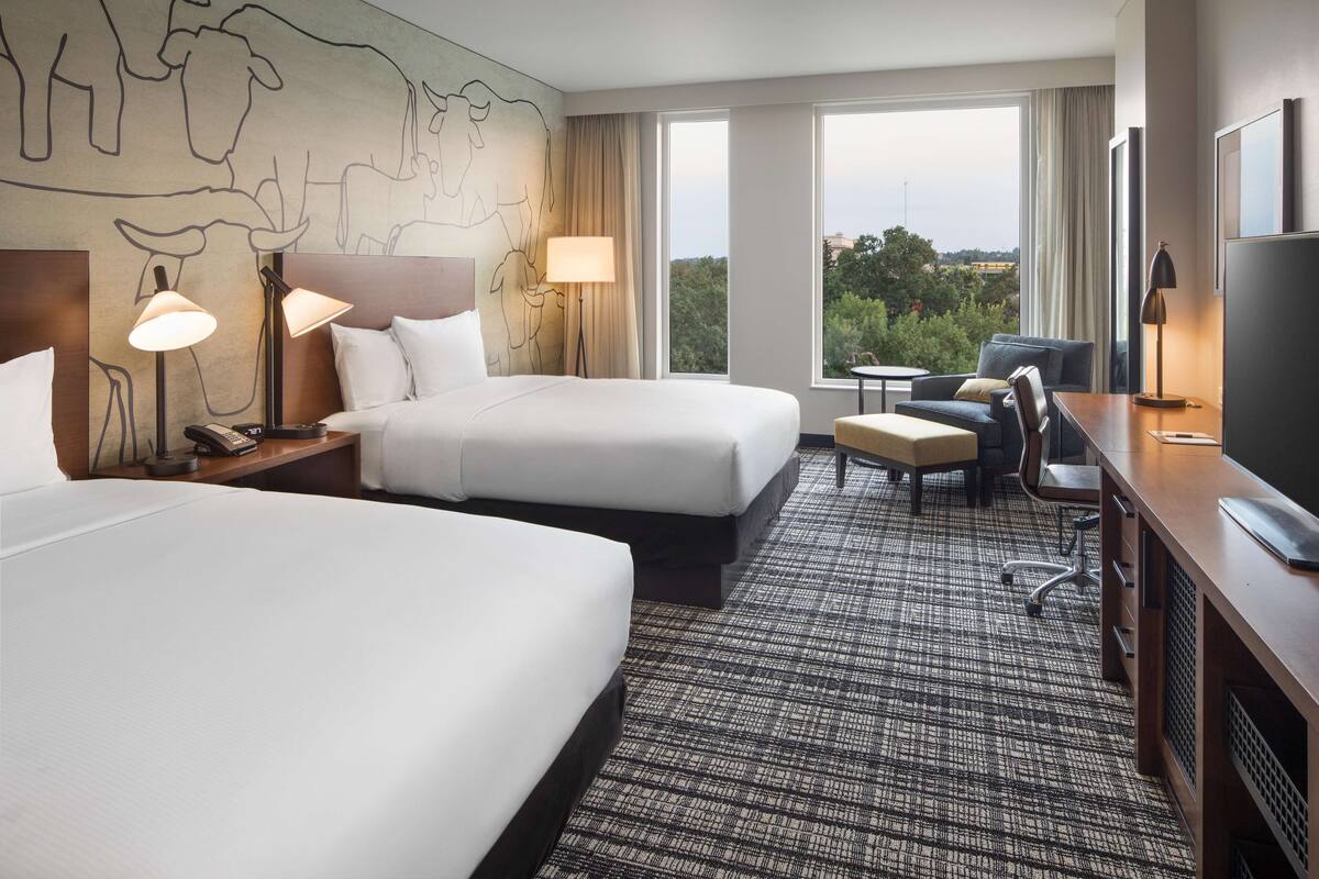 A hotel room with two beds, a desk with a TV, a chair, a lamp, and a view of the outdoors.