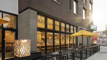 An outdoor cafe with tables and chairs, a brick building facade, and a large yellow umbrella.