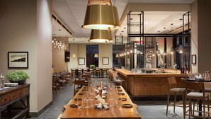 A modern dining area with a long wooden table set for a meal, surrounded by chairs, and a bar area with stools and a counter. - DoubleTree by Hilton Greeley at Lincoln Park (Greeley)