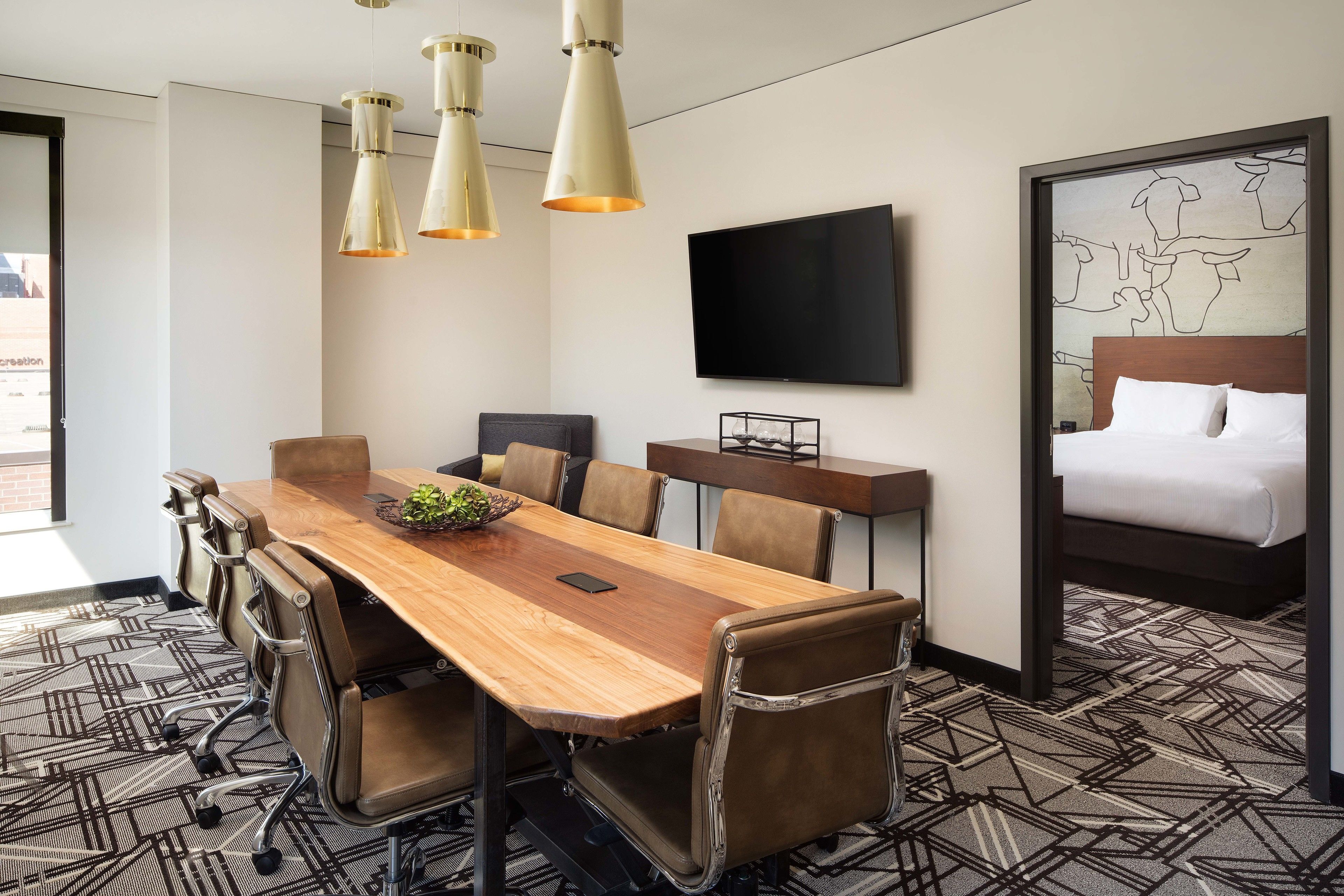 A hotel room with a wooden conference table, leather chairs, a bed with a headboard, a flat-screen TV mounted on the wall, and a patterned carpet.