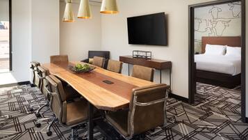 A hotel room with a wooden conference table, leather chairs, a bed with a headboard, a flat-screen TV mounted on the wall, and a patterned carpet.