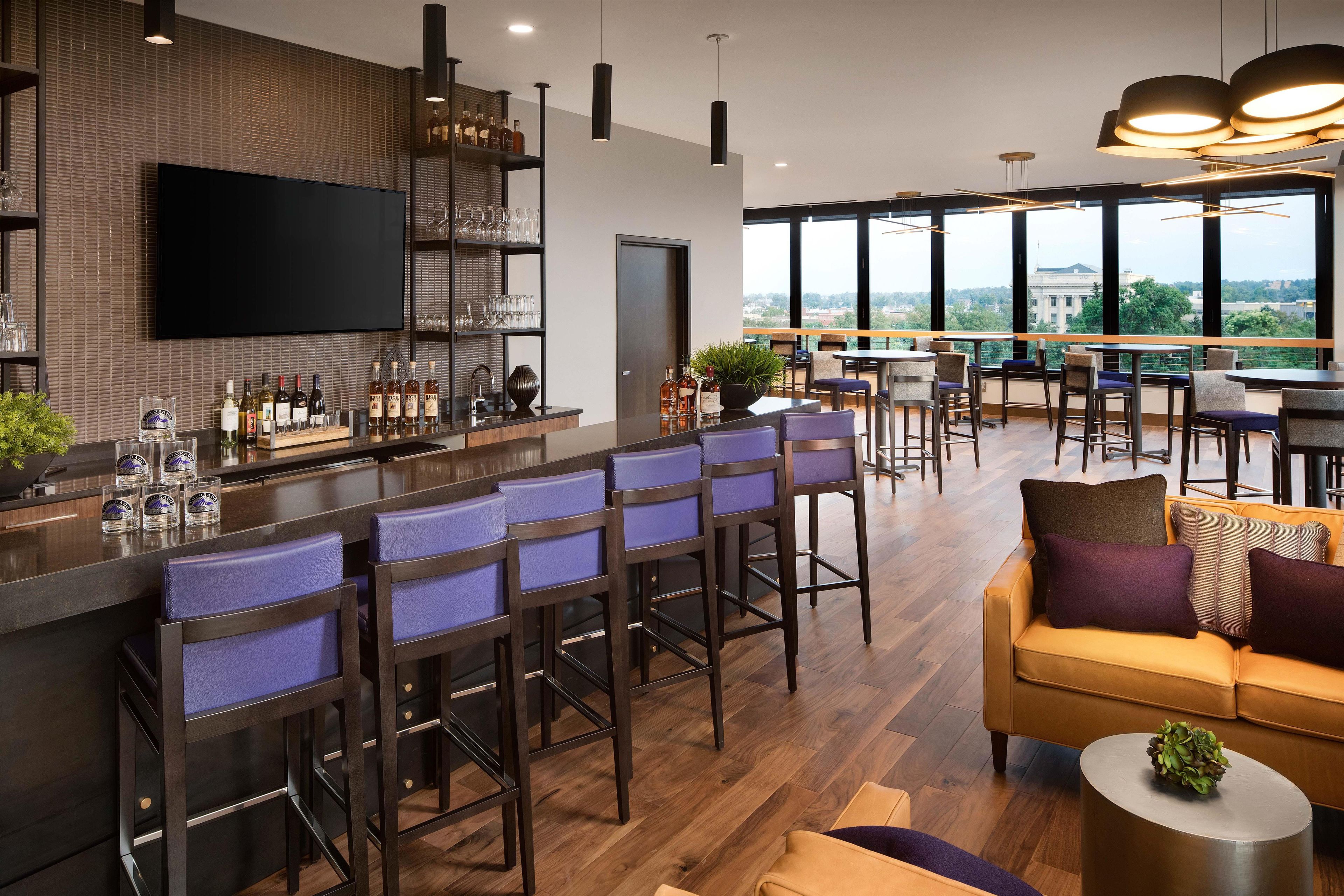 A modern bar area with high stools, a flat-screen TV, and a view of the city through large windows.