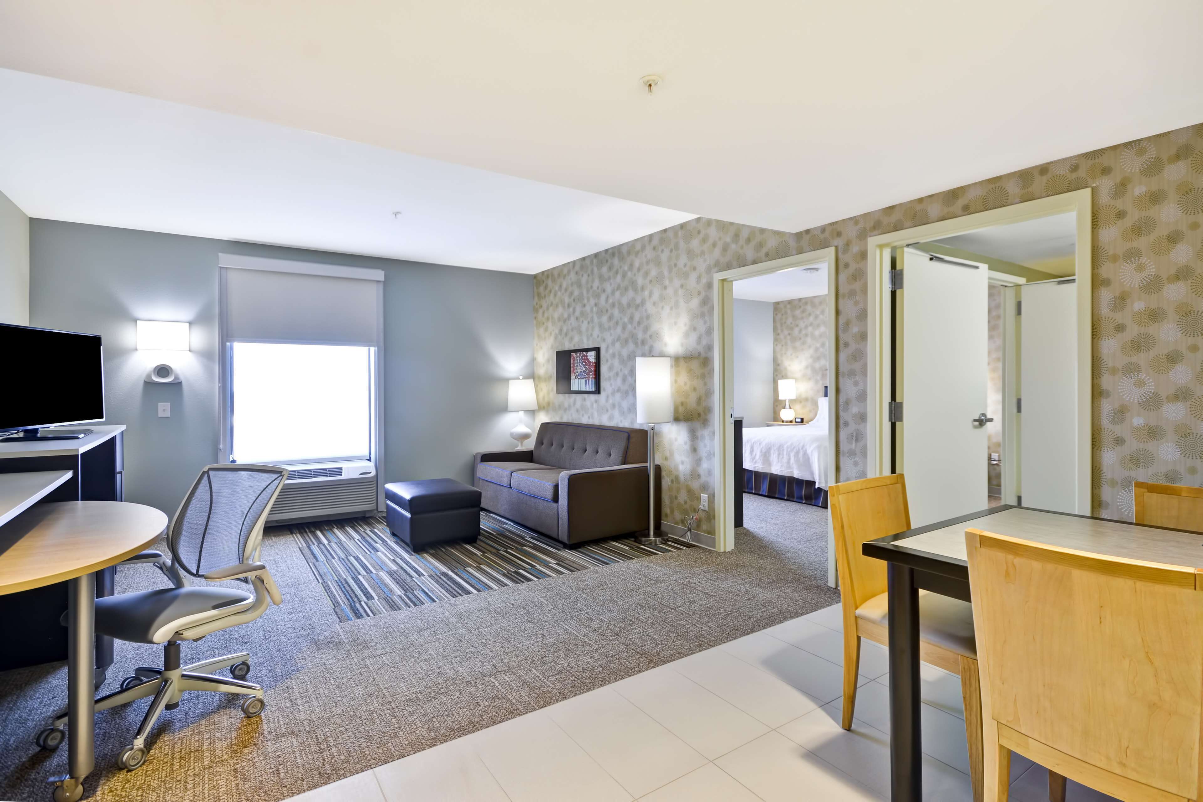 A hotel room with a desk, office chair, sofa, and a dining table. There are two lamps, a TV, and a bed in the background.