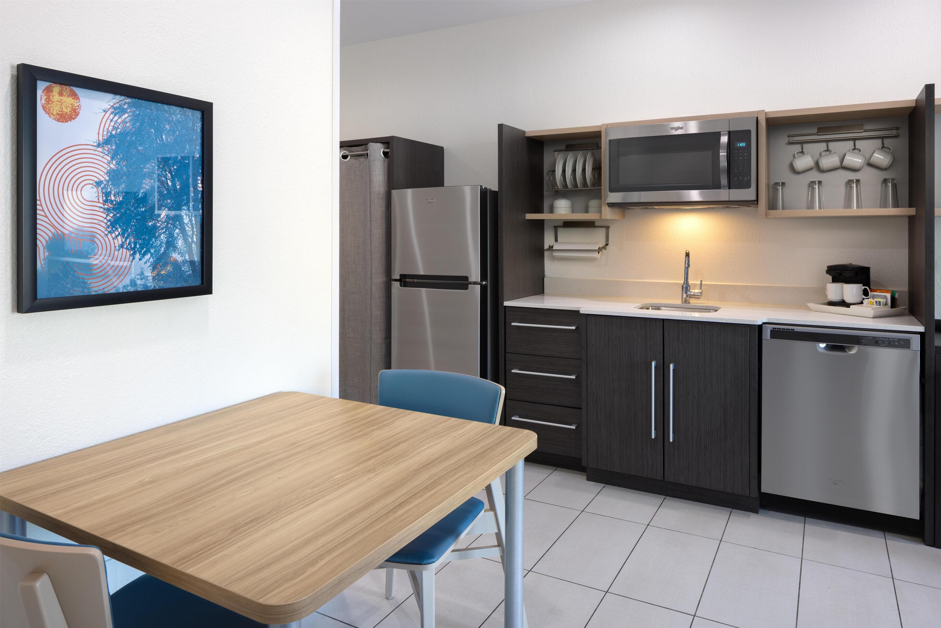 Een moderne keuken met een roestvrijstalen koelkast, magnetron en vaatwasser, een houten eettafel en een blauwe stoel.