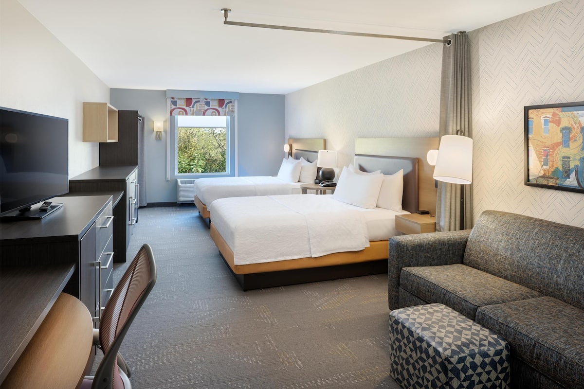 A hotel room with a large bed, a sofa, a desk with a TV, and a window with a view of greenery.