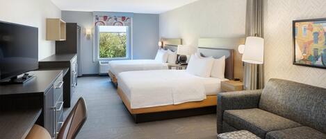 A hotel room with a large bed, a sofa, a desk with a TV, and a window with a view of greenery.