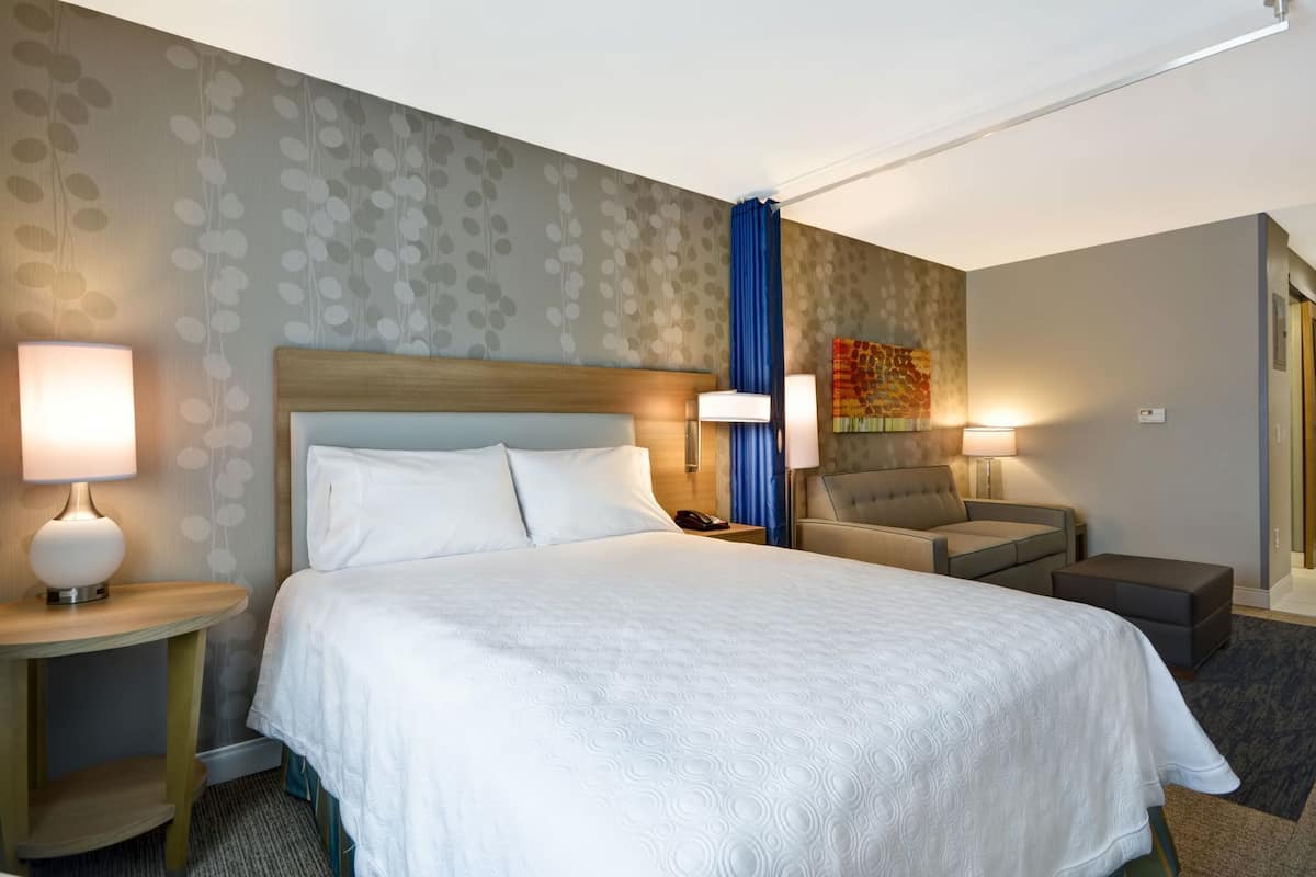 A hotel room with a large bed, two bedside tables with lamps, a chair, and a small ottoman.