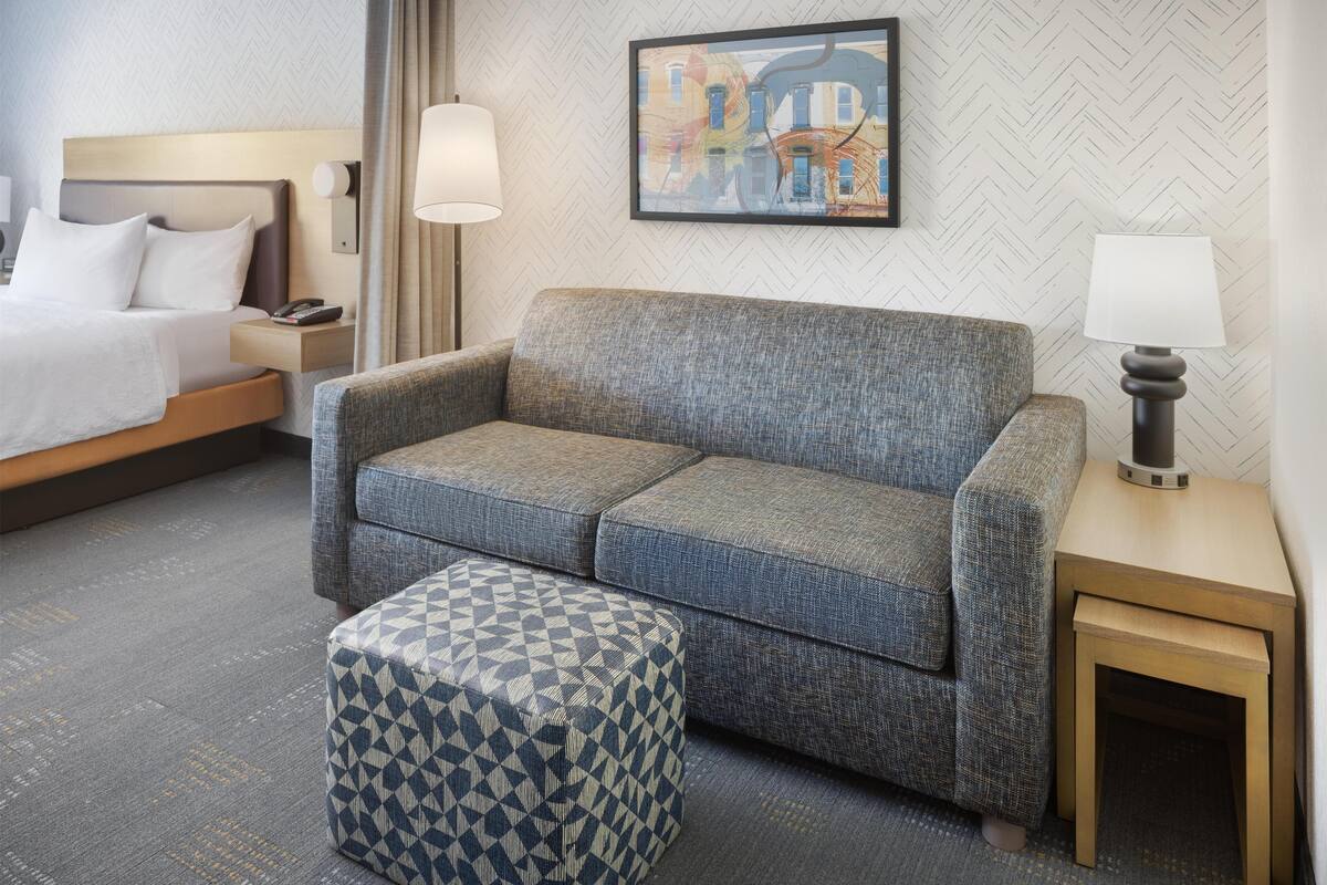 A hotel room with a bed, a grey sofa, a patterned ottoman, a small wooden side table, and a wall-mounted lamp.