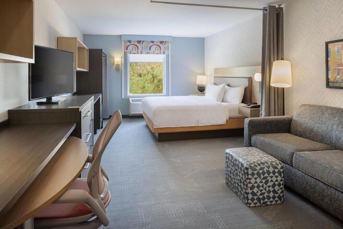 A hotel room with a bed, a desk with a TV, a sofa, and a window with a view.