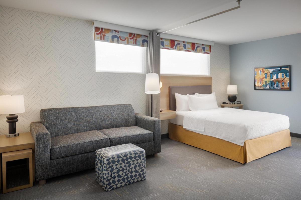 A hotel room with a grey sofa, a patterned ottoman, a bed with white bedding, a nightstand with a lamp, and a wall with a colorful abstract painting.