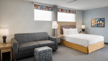 A hotel room with a grey sofa, a patterned ottoman, a bed with white bedding, a nightstand with a lamp, and a wall with a colorful abstract painting.