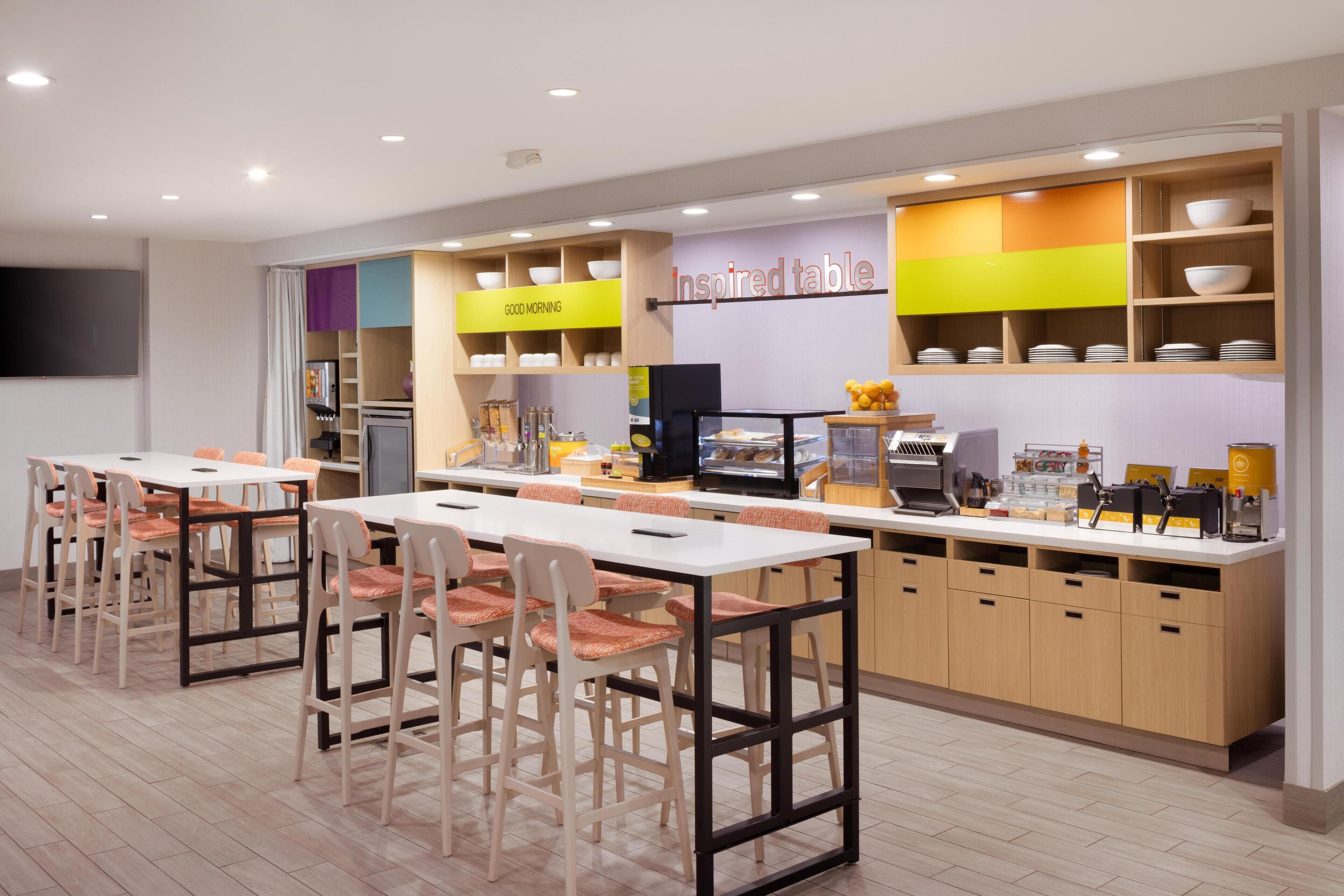 A modern kitchen area with multiple tables and chairs, a counter with various kitchen appliances, and shelves with bowls and dishes.