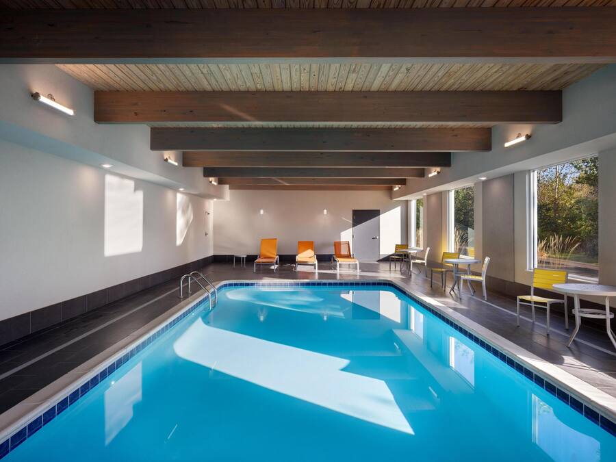 A modern indoor swimming pool with clear blue water, wooden ceiling beams, and seating areas with chairs and tables.