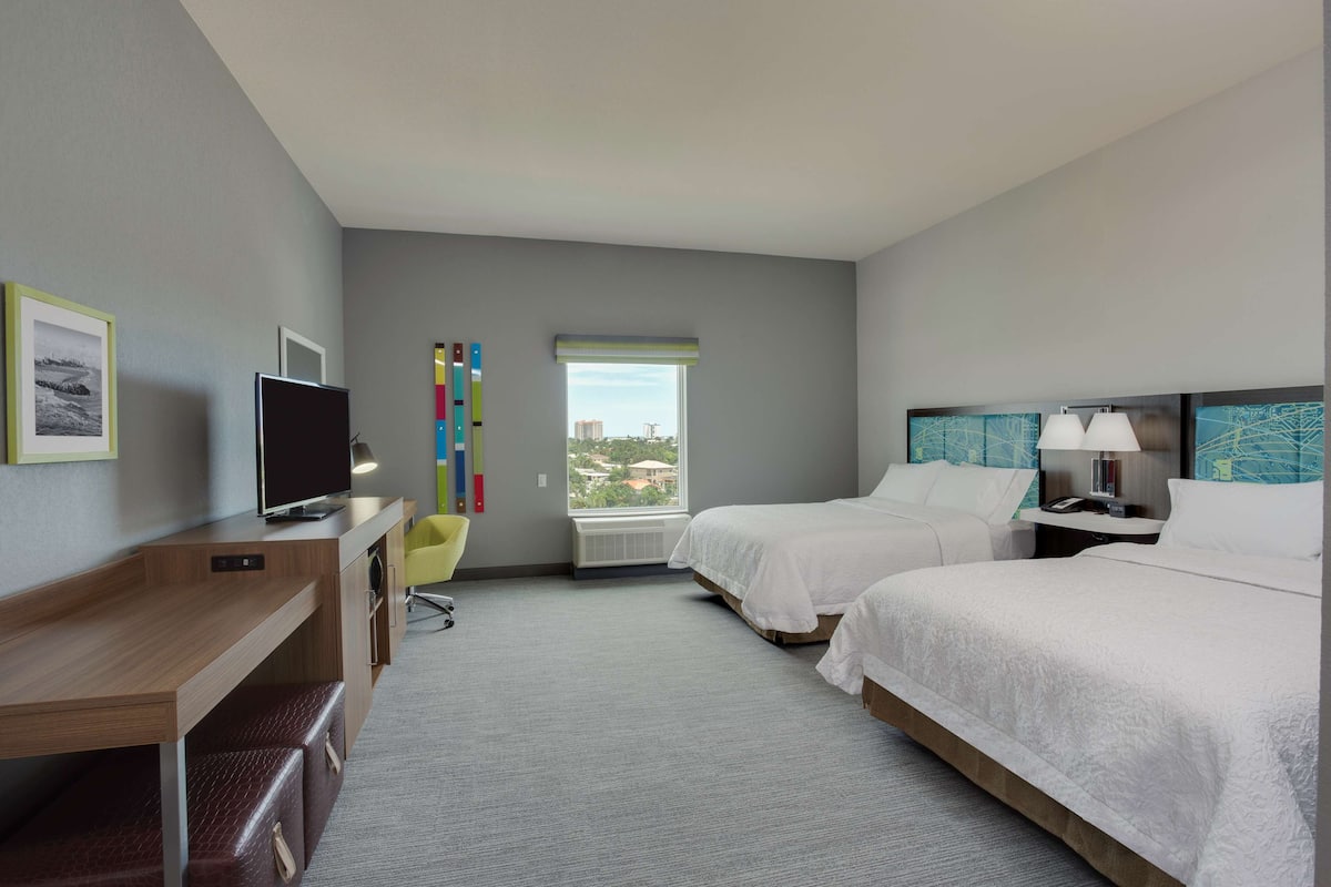 A hotel room with two beds, a desk, a TV, a chair, and a window with a view.