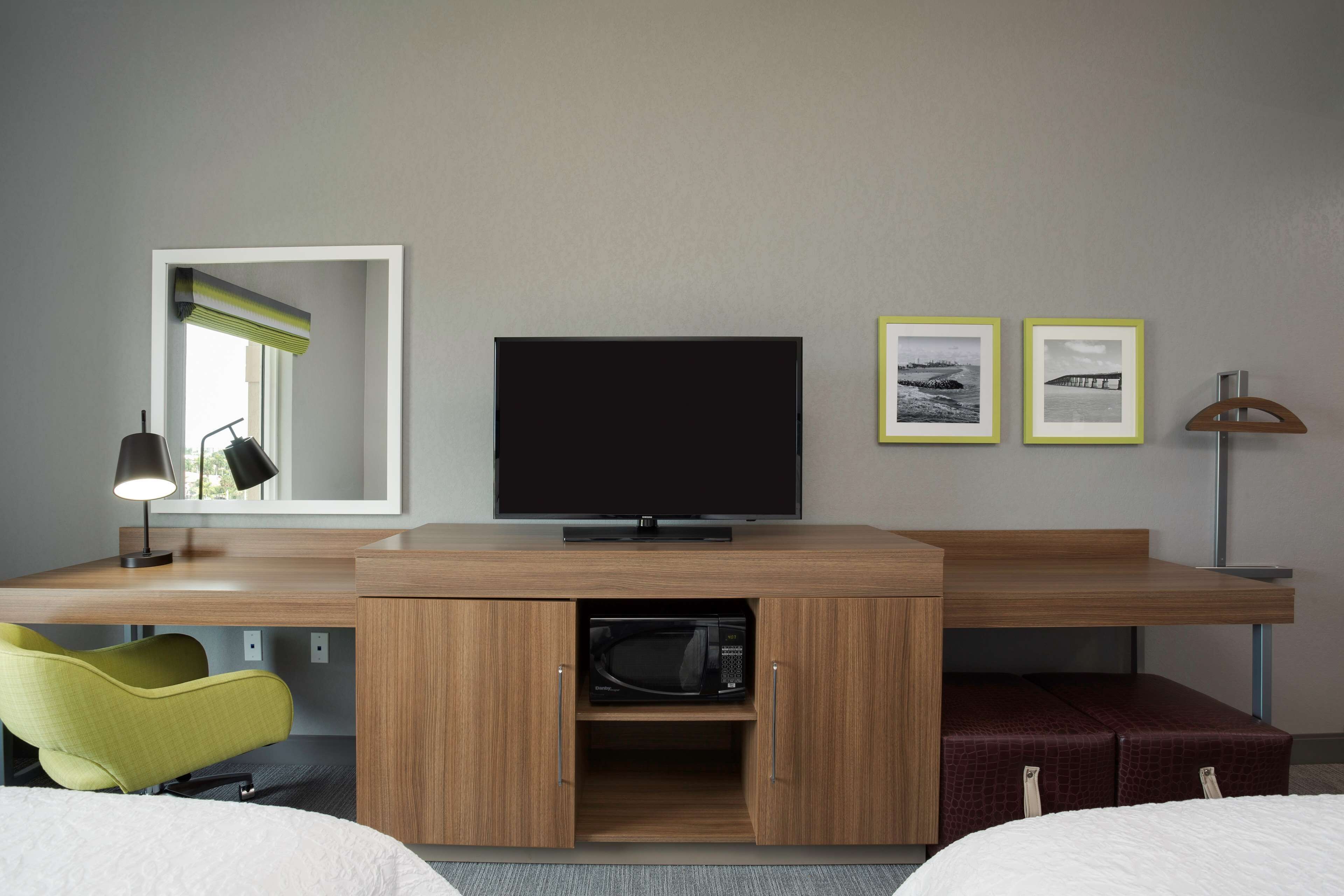 A hotel room with a wooden desk, a television, a microwave, a green chair, and two framed pictures on the wall.