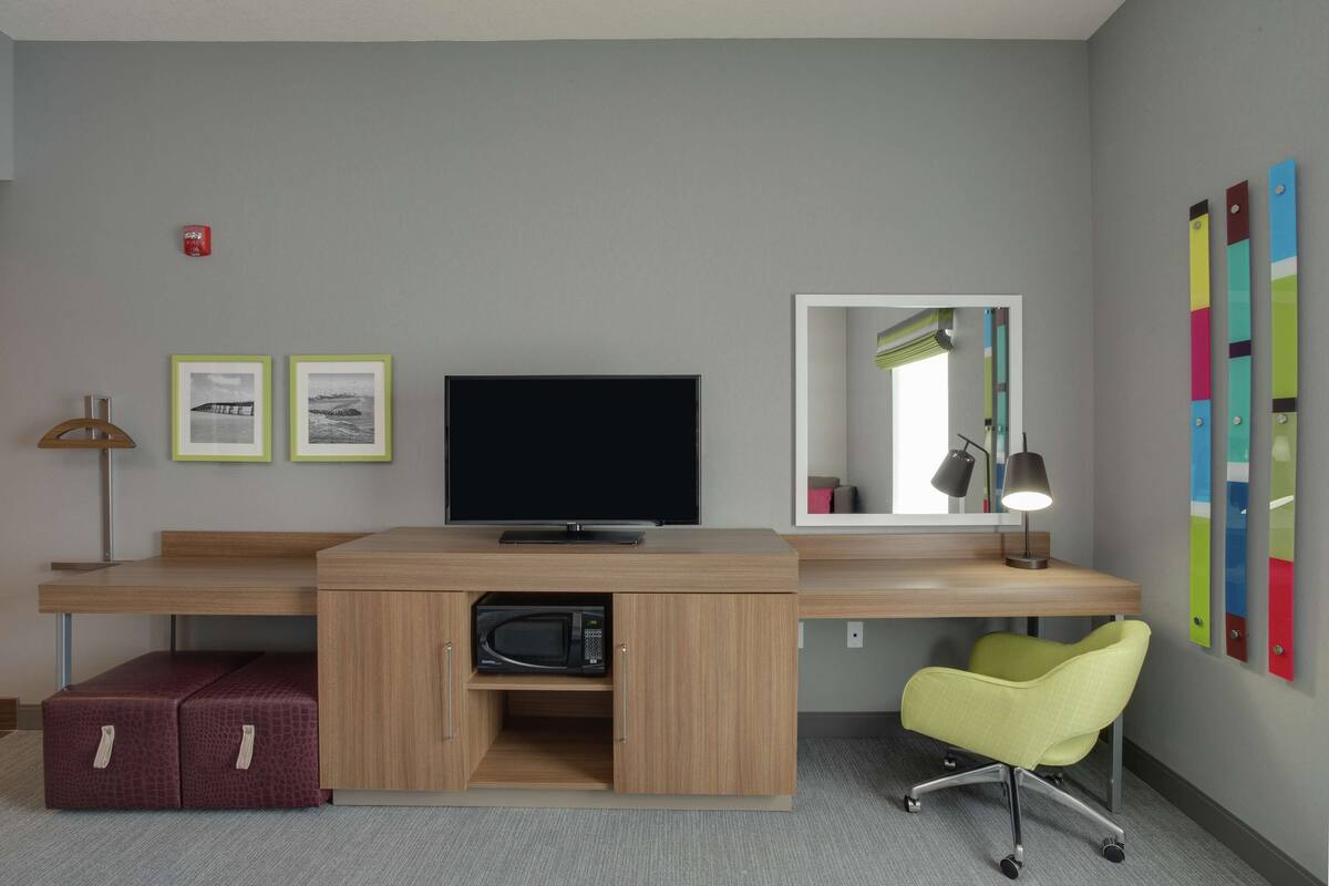 A hotel room with a wooden desk, a television, a microwave, a yellow chair, and a mirror.