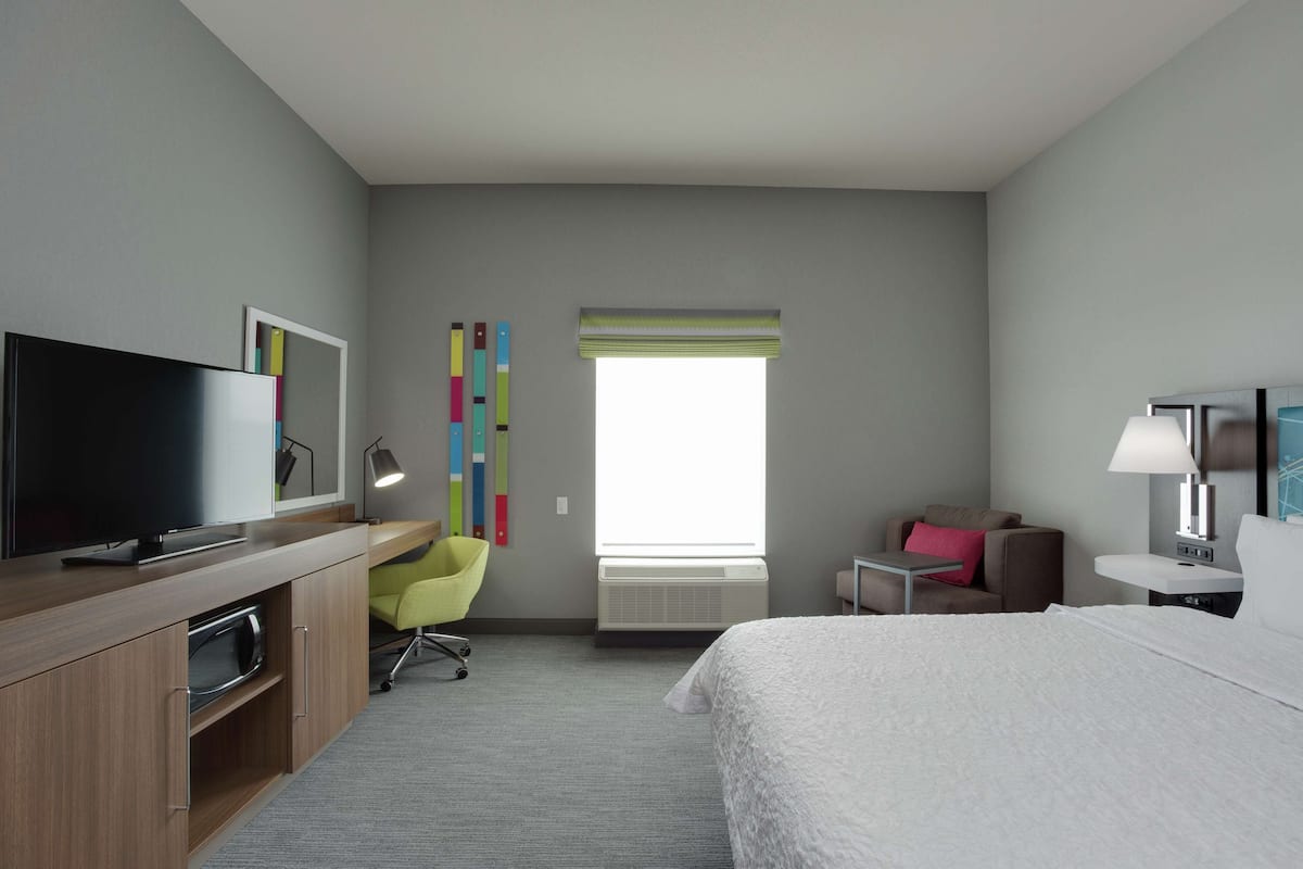 A hotel room with a bed, a desk with a TV, a chair, a small table, and a window with blinds.