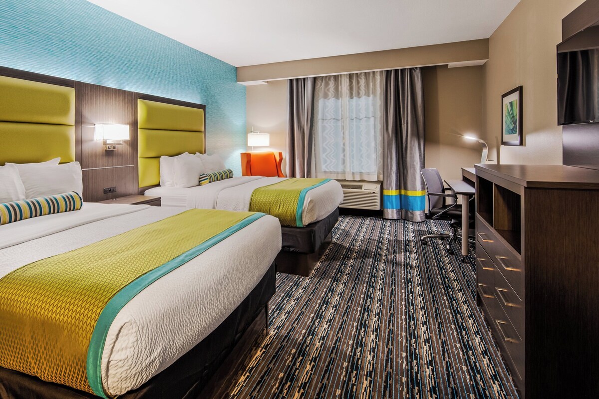 A hotel room with two beds, a desk, a chair, a TV, and a large window with curtains.