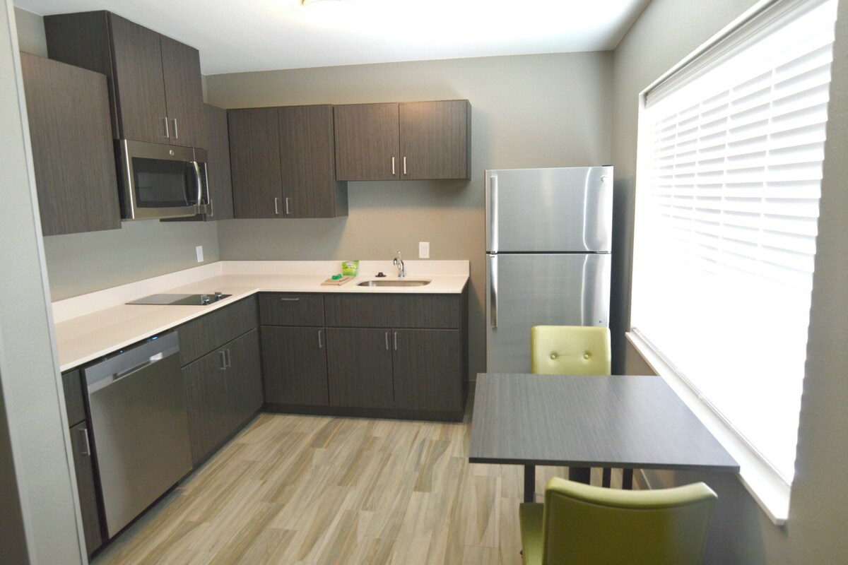A compact kitchen with dark cabinets, a stainless steel refrigerator, a microwave, and a dishwasher. There is a small dining table with two chairs.