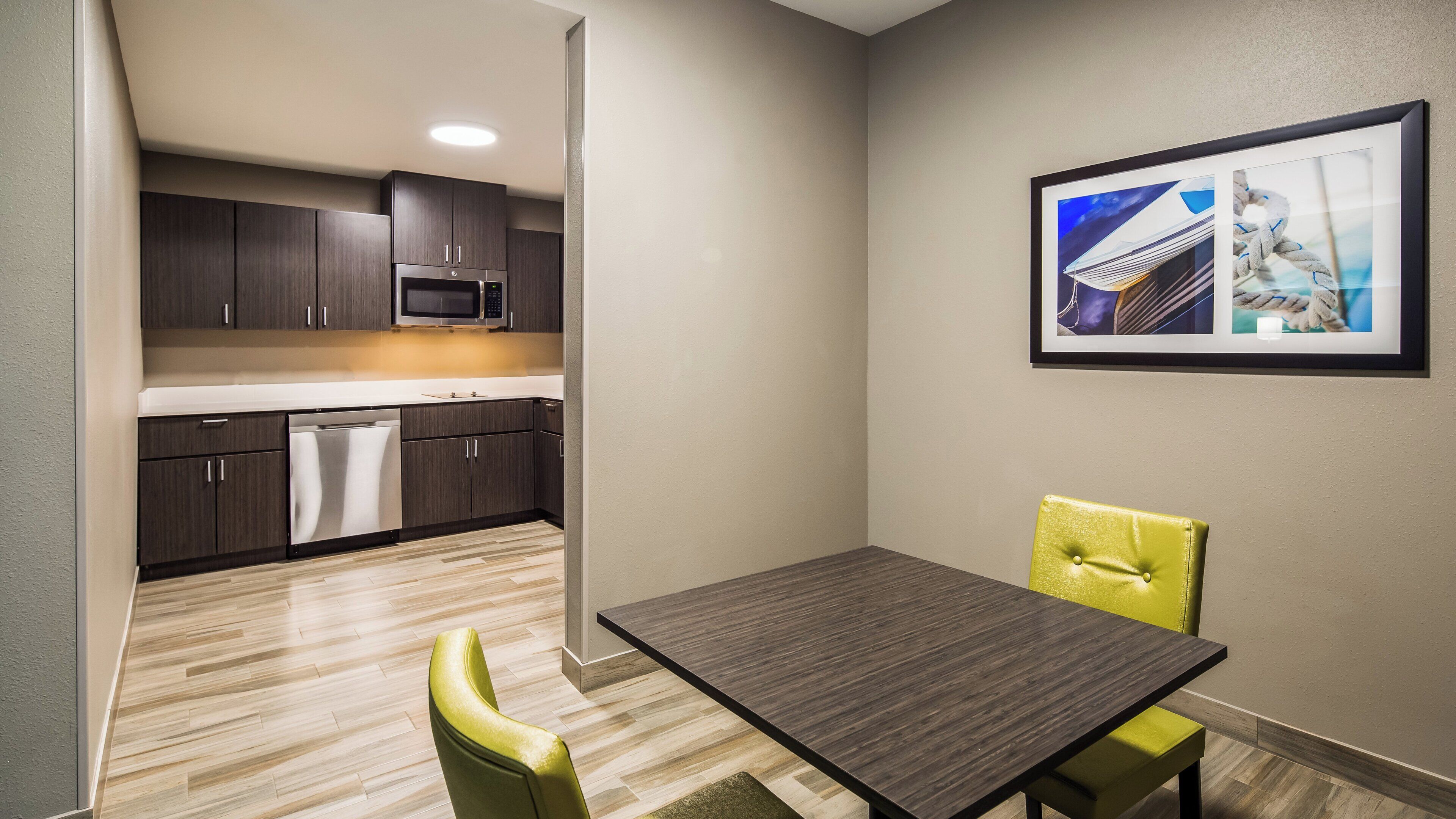 A compact kitchen with dark cabinets, a microwave, and a dishwasher. There is a dining table with two chairs and a framed picture on the wall.
