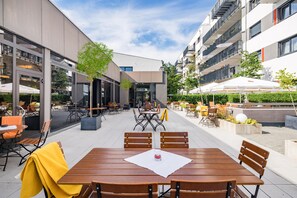 An outdoor seating area with tables and chairs, surrounded by buildings and greenery.